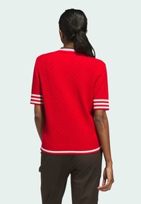 Pull rouge à manches courtes avec un motif texturé, des poignets rayés blancs et rouges, et un col contrastant. Coupe simple et texture en tricot lisse.