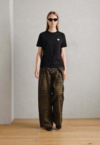 T-shirt nera con logo sul petto; pantaloni larghi con stampa leopardata in marrone e nero; il modello indossa occhiali da sole scuri; sfondo neutro.