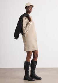 Beige Sweatshirtkleid mit abgerundetem Saum, weißen Bündchen und seitlichen Taschen. Schwarze knielange Stiefel. Helle beige Mütze mit Aufschrift. Schwarze Jacke lässig über die Schulter gelegt.