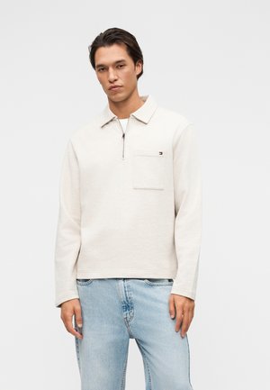 Νέος άνδρας που φοράει ένα κρεμ πουλόβερ με μακριά μανίκια και μισό φερμουάρ στο στήθος με τσέπη, συνδυασμένο με ανοιχτό μπλε τζιν, στέκεται μπροστά σε απλό φόντο.