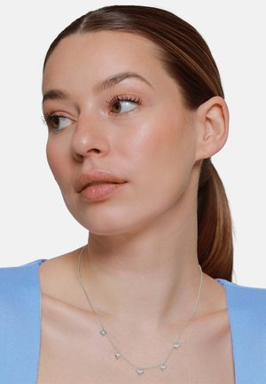 Donna con capelli castani raccolti all'indietro che indossa una maglietta azzurro chiaro e una collana d'argento con piccoli ciondoli a forma di farfalla, guarda verso sinistra.