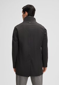 Cappotto nero con colletto alto e taglio sartoriale, caratterizzato da uno spacco sul retro e una texture liscia e opaca. Pantaloni grigi indossati sotto.