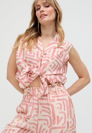 Frau trägt ein ärmelloses, geknöpftes Hemd, das an der Taille gebunden ist, und passende, hoch taillierte Hosen mit rosa abstrakten Mustern auf beigem Hintergrund.