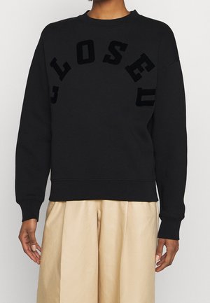 Personne portant un sweat-shirt noir avec des lettres noires en relief formant "CLOSED" en arc sur la poitrine et un pantalon beige à larges jambes.