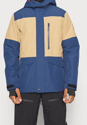 Mann trägt eine blaue und beige Kapuzen-Winterjacke mit Reißverschlusstasche auf der Brust und schwarze Schneehose mit Reißverschlussdetails.