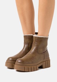 Brązowe skórzane botki za kostkę o teksturowanym wykończeniu, z puszystym kremowym wykończeniem przy kołnierzu i masywną, żebrowaną gumową podeszwą.