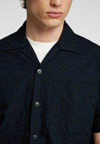 Košile s krátkým rukávem v tmavě modré barvě, s vyšívaným vzorem, náprsní kapsou a knoflíkovým zapínáním. Měkká textura s viditelnými dvěma knoflíky.