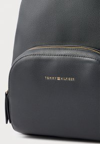 Černý kožený batoh s texturovaným povrchem, vybavený zlatým zipem a zlatým logem na přední straně. Zaoblený tvar s elegantním designem.