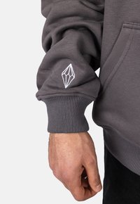 Felpa grigia con polsini a coste, caratterizzata da un logo geometrico ricamato bianco sul maniche sinistro. Il tessuto appare morbido e dalla consistenza piacevole.