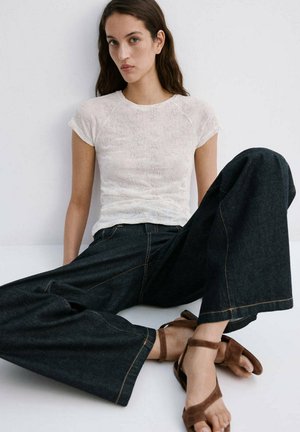 Jeune femme assise contre un mur blanc, portant un haut blanc texturé à manches courtes, un jean en denim foncé à jambes larges et des sandales marron à brides.