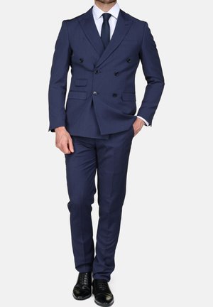 Man draagt een marineblauw dubbelrijks colbert met bijpassende broek, wit overhemd, zwarte das en zwarte nette schoenen, staand met één hand in de zak.