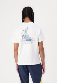 Hvit bomull t-skjorte med et blått grafisk trykk av en fiskebåt på baksiden, med "New Balance" tekst og omkransende fugler.