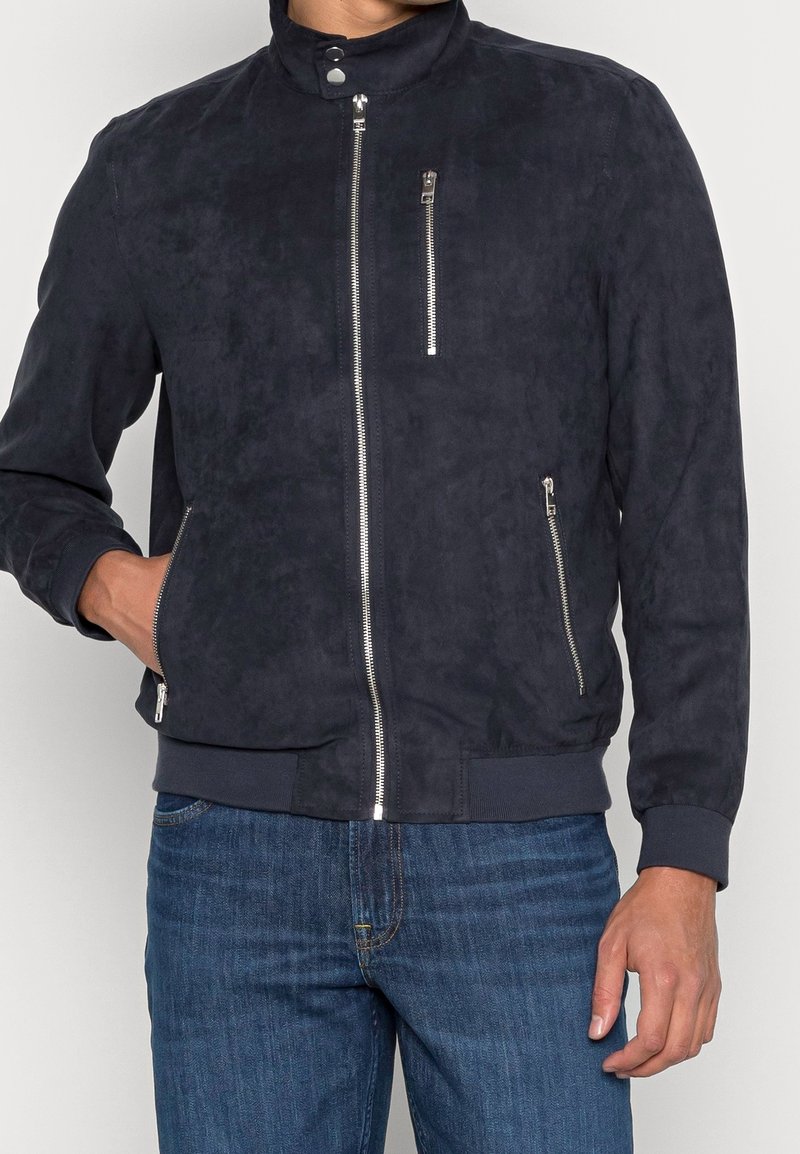 Marynarka bomberka zamszowa w kolorze granatowym, z kołnierzem stójką, przednim suwakiem i dwoma bocznymi kieszeniami; ściągacze na mankietach i u dołu dla dopasowanego wykończenia.