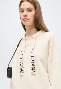 Jeune femme aux cheveux châtain clair lisses portant un sweat à capuche crème avec des cordons à motifs noirs et un petit sac à bandoulière noir.