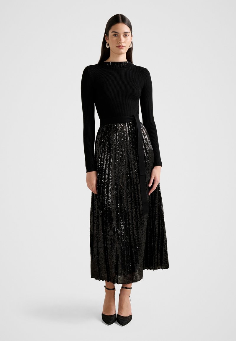 Mujer que lleva un top negro de manga larga con cuello alto y una falda midi plisada negra con lentejuelas, combinada con tacones y pendientes de aro.