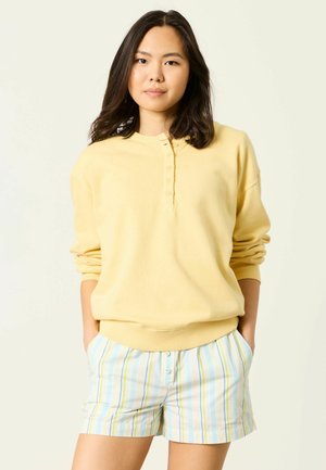 Mladá žena s tmavými vlasy, která má na sobě žluté tričko s dlouhým rukávem a proužkované pastelové šortky, stojí s rukama v kapsách na světlejší zázemí.