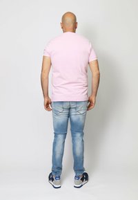 T-shirt di cotone rosa pallido, maniche corte, indossata con jeans in denim blu chiaro e sneakers nere. Cuciture visibili e due tasche posteriori sui jeans.