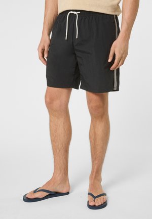 Uomo che indossa pantaloncini neri con coulisse e strisce laterali bianche, e infradito blu navy, in piedi contro uno sfondo uniforme.