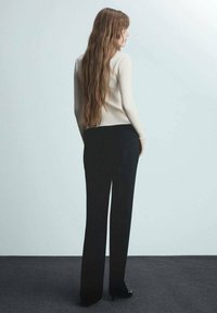 Maglione a coste crema e pantaloni neri a gamba dritta. Lo sfondo è grigio chiaro, creando contrasto con l'abbigliamento e presenta texture morbide.