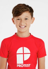 Raudonas rash guard'as su trumpomis rankovėmis, kuris turi didelį baltą grafikos logotipą priekyje. Medžiaga atrodo elastinga ir lygi.