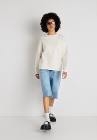 Světle béžová mikina s textura rukávy, doplněná světlými modrými džíny šortkami a černými dřeváky. Model má na sobě průhledné sluneční brýle a bílé ponožky.