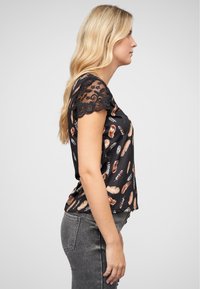 Chemise noire à manches courtes avec un motif de plumes de différentes couleurs, ornée de dentelle sur les manches et d'une coupe décontractée.