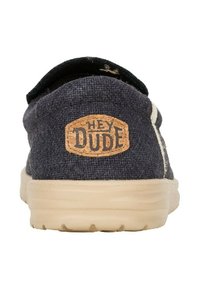 Zapato slip-on de lona azul marino con suela de goma beige. Presenta una etiqueta de corcho que dice "HEY DUDE" en la parte posterior. Diseño simple, sin patronesintrincados.