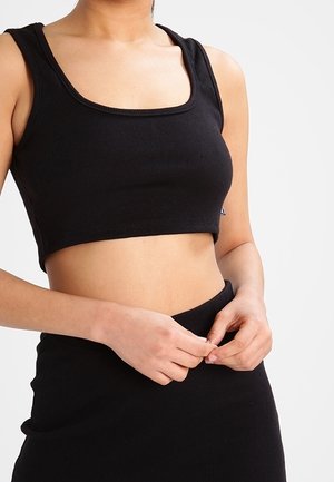 Kvinde iført sort ribbet crop top og matchende højtaljet nederdel, hænder justerer nederdelen ved taljen mod en lys baggrund.