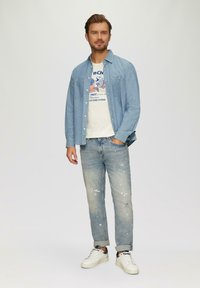 Lichtblauw denim shirt over een wit graphic T-shirt, gecombineerd met versleten lichtblauwe denim jeans en witte sneakers.