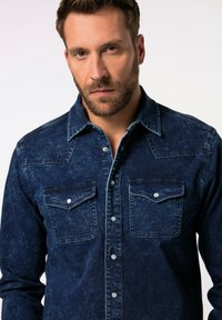 Mørkeblå denimskjorte med små brystlommer, trykknapper, spids yoke-design og en falmet struktur. Klassisk western-stil.
