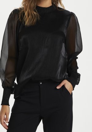 Femme portant un chemisier noir avec des manches bouffantes transparentes et des poignets froncés, associée à un pantalon noir, une main dans la poche.