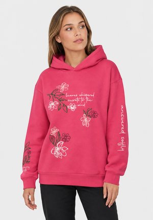 Vrouw die een roze hoodie draagt met bloemmotieven en witte tekst met daarop "leaves whispered secrets to the wind" en "lotus pardansana" op de mouw.