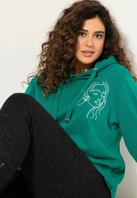 Felpa color teal con design di volto ricamato bianco, dotata di lacci e punte in metallo. Abbinata a pantaloni neri strutturati. Tessuto morbido, vestibilità casual.