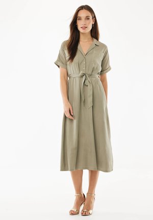 Kvinde med langt brunt hår iført en khaki midi-kjole med knapper, korte ærmer og et bundet bælte i taljen, stående i gyldne åbne stiletter.