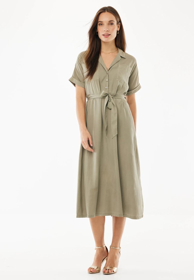 Frau mit langen braunen Haaren, die ein khakifarbenes knielanges Hemdkleid mit kurzen Ärmeln und einem gebundenen Taillengürtel trägt, steht in goldfarbenen offenen High-Heels.
