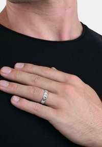 Anello d'argento con motivo geometrico, forma rotonda, superficie liscia; indossato su una mano con tono di pelle neutro, su sfondo di camicia nera.