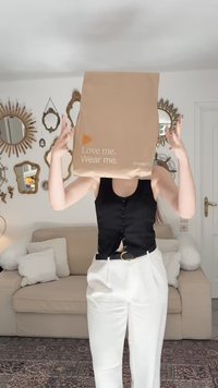 Sac en papier marron portant le texte "Aime-moi. Porte-moi." et le logo Zalando, tenu au-dessus d'une personne portant un haut sans manches noir et un pantalon blanc.