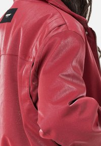 Rode jas van imitatieleren met een gladde textuur, oversized pasvorm en een zwart label op de bovenrug. Ritssluiting detail zichtbaar.