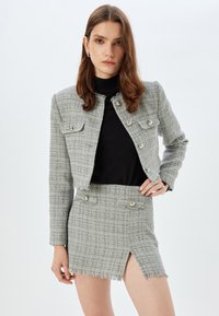Giacca cropped in tweed grigio con bordi sfrangiati e due tasche frontali, abbinata a una mini gonna in coordinato con uno spacco laterale e due bottoni.