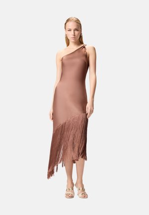 Kvinde stående iført en ærmeløs, enkeltskulder brun kjole med asymmetrisk frynsekant og beige åbne hæle med guld accenter.