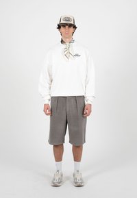 Homme portant un sweat-shirt blanc, un short gris jusqu'aux genoux, des baskets blanches, une casquette beige et un foulard à motifs autour du cou, debout devant un fond uni.