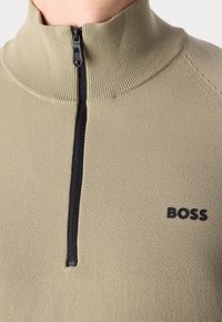 Beige texturerad stickad tröja med svart kvartslång dragkedja och svart "BOSS"-logotyp på vänster bröst.