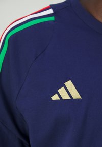 Granatowa koszulka z wielokolorowymi paskami w czerwonym, białym i zielonym kolorze na ramionach. Złota haftowana logo Adidas po lewej stronie klatki piersiowej.