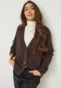 Cardigan texturé marron foncé avec un col en V, fermeture à boutons et poignets côtelés, porté sur un pull col roulé crème et un pantalon noir.