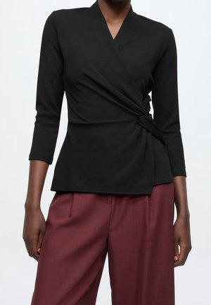 Femme portant un top noir à manches longues avec un nœud sur le côté et un pantalon bordeaux ample sur fond uni.