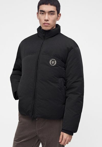 Jeune homme portant une doudoune noire zippée avec un patch logo rond sur la poitrine et un pantalon en velours côtelé marron.