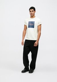 Cream-colored t-shirt with a blue square graphic reading "BARBOUR INTERNATIONAL." Paired with loose black pants and black shoes.