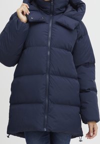 Námořnický modrý péřový kabát s vysokým límcem, zapínáním na zip a třemi vodorovnými prošívanými sekcemi. Obsahuje odnímatelnou kapuci a elastické manžety.