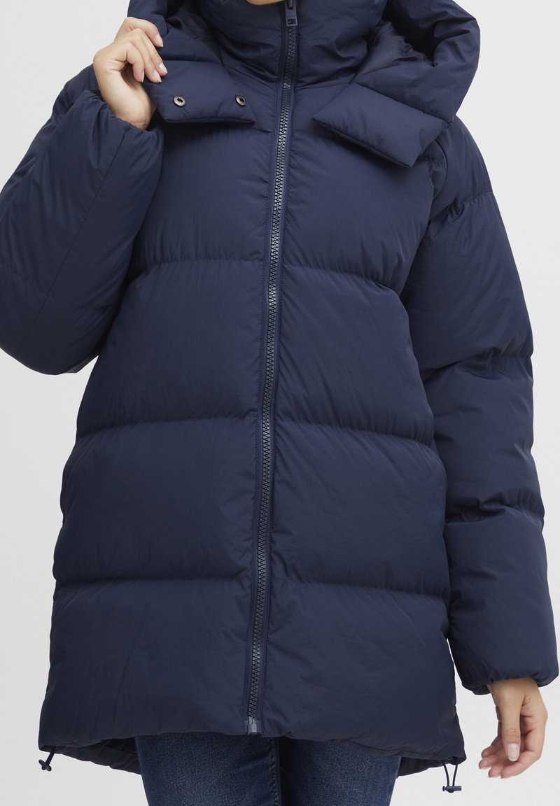 Námořnický modrý péřový kabát s vysokým límcem, zapínáním na zip a třemi vodorovnými prošívanými sekcemi. Obsahuje odnímatelnou kapuci a elastické manžety.