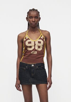 Žena s vrkočmi, ktorá má na sebe hnedý top na viazanie s číslom "98" a čiernu rifľovú minisukňu na jednofarebnom pozadí.